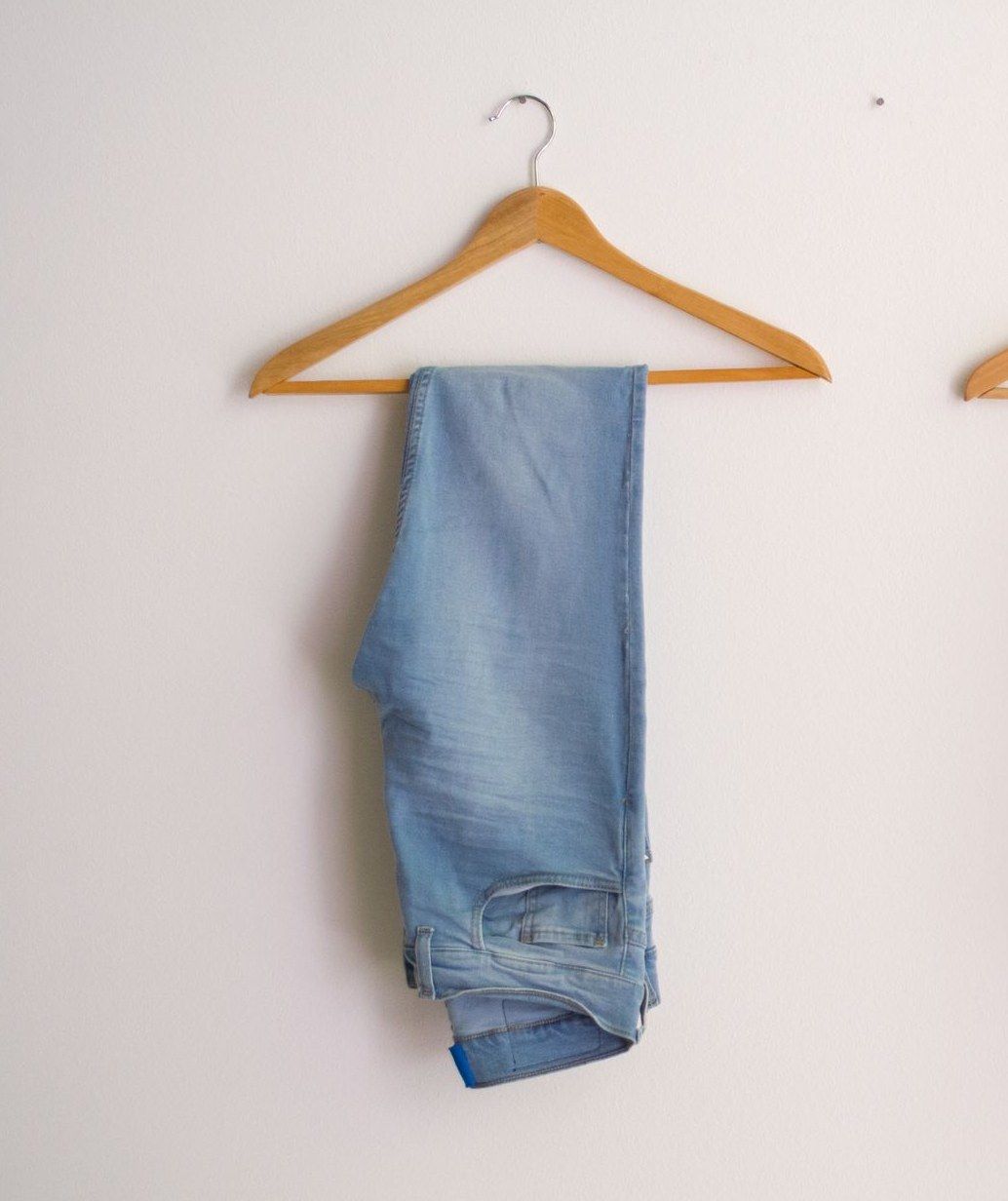 Eine hellblaue Jeans hängt gefaltet über einem Holzbügel an einer weißen Wand